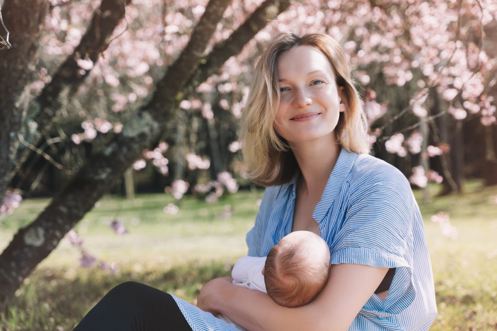 En ung kvinne sitter på en gressplen, og ser i kameraet og smiler mens hun ammer et barn. Foto
